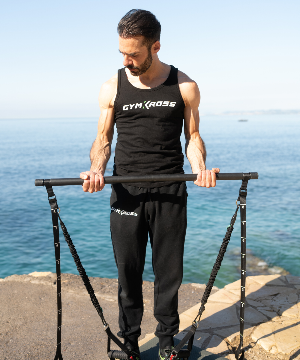 Un homme en débardeur noir et pantalon "GYMKROSS" utilise des bandes de résistance attachées à une barre sur une promenade côtière. La mer bleue est visible en arrière-plan sous un ciel dégagé.