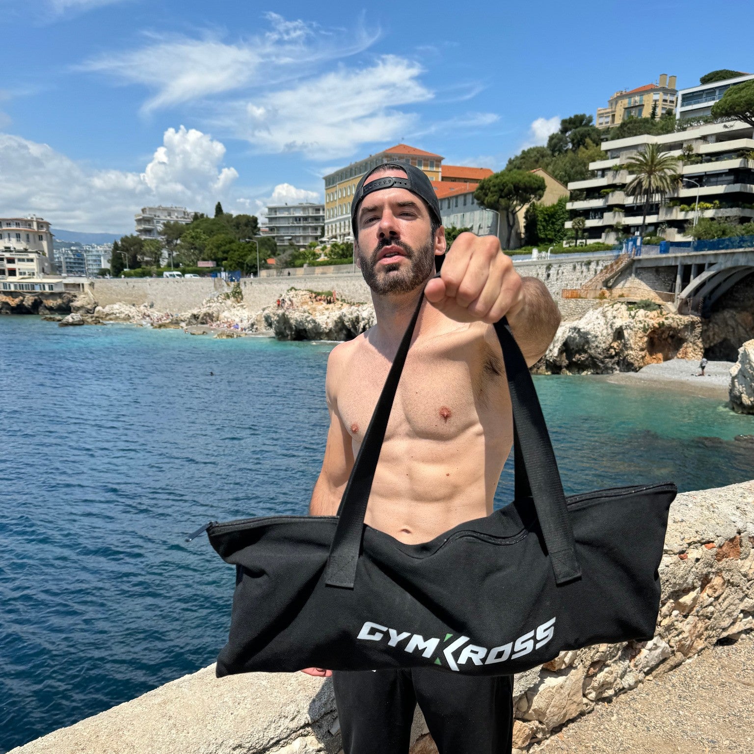 Un homme torse nu portant un pantalon noir et une casquette à l'envers, tient un sac de sport noir marqué "GYMKROSS" sur une promenade côtière. En arrière-plan, on voit une plage rocheuse, des bâtiments et une mer bleue sous un ciel partiellement nuageux.