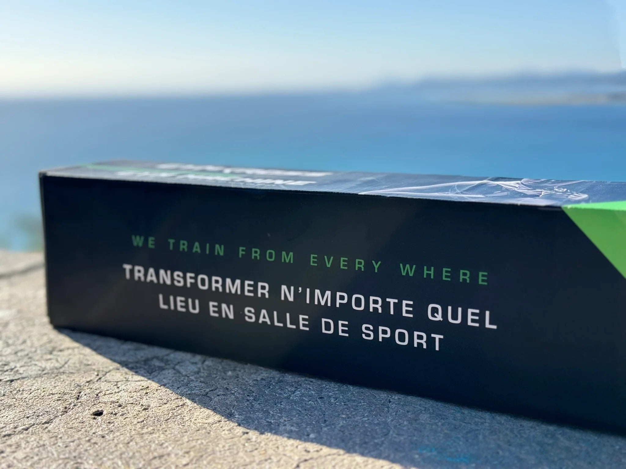 Boîte GymKross avec le slogan 'WE TRAIN FROM EVERY WHERE - TRANSFORMER N’IMPORTE QUEL LIEU EN SALLE DE SPORT', posée sur un muret avec la mer en arrière-plan, suggérant la liberté d'entraînement n'importe où avec GYM-KROSS.