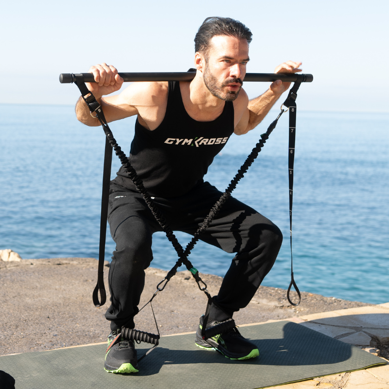 &quot;Un homme en tenue de sport GymKross effectuant des squats avec des élastiques et une barre au bord de l&#39;eau. L&#39;entraînement en plein air avec GymXross permet de renforcer les muscles et d&#39;améliorer la santé tout en profitant des bienfaits du soleil et de l&#39;exercice en extérieur.&quot;