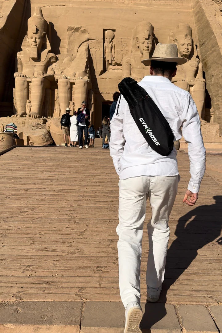 Un voyageur en tenue décontractée blanche et chapeau, dos tourné à la caméra, marchant vers le temple d'Abou Simbel en Égypte, équipé d'un sac de sport GymKross. Cette image illustre l'union du voyage et du fitness, soulignant comment GymKross facilite le maintien d'une routine d'entraînement même lors de la visite de sites emblématiques mondiaux.