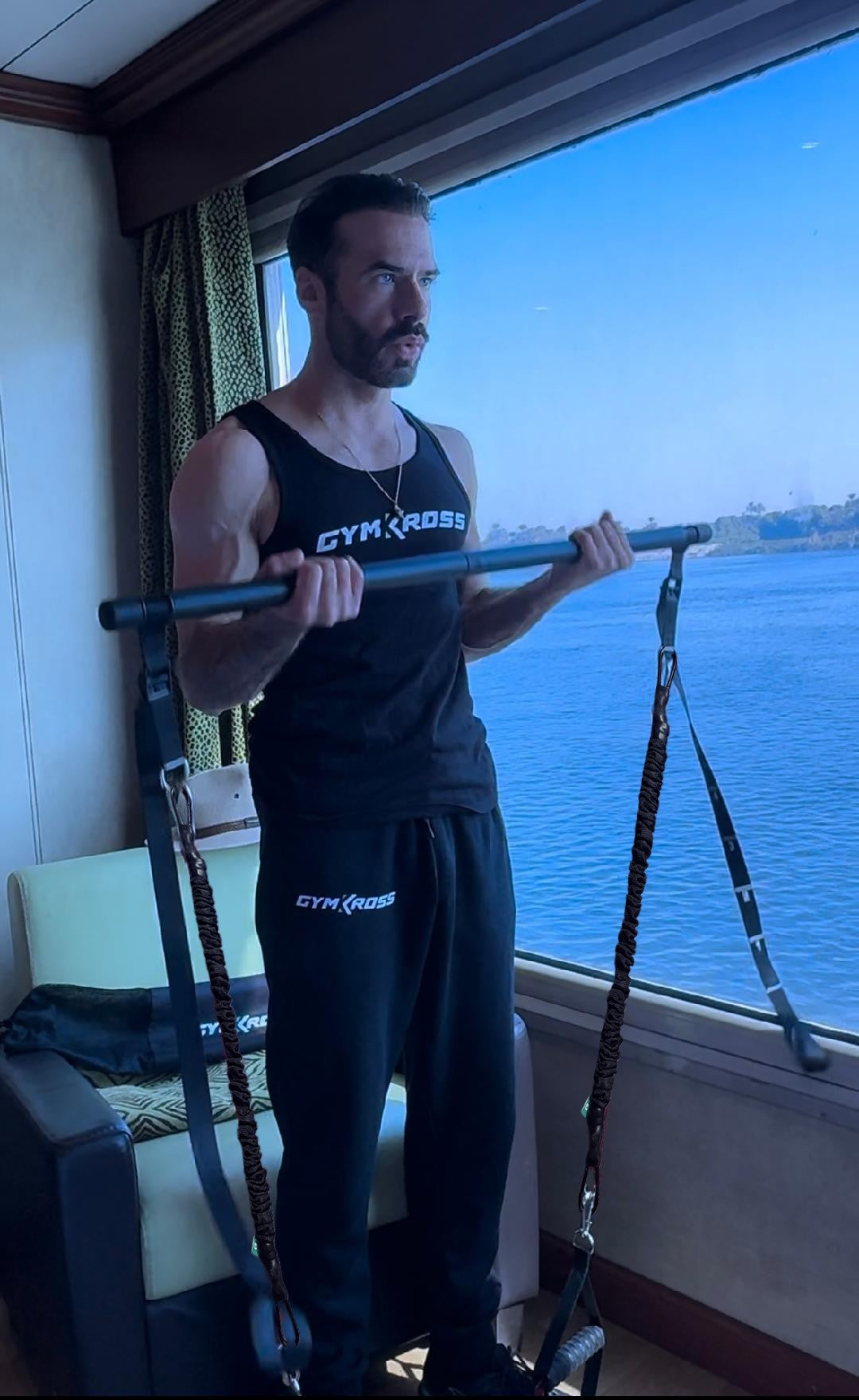 Un homme s'exerce avec des bandes de résistance dans une pièce avec une grande fenêtre offrant une vue panoramique sur une étendue d'eau et un paysage lointain. Il porte un débardeur noir et un pantalon "GYMKROSS".