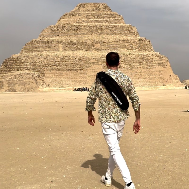 Un homme marche vers la Pyramide de Djoser, une ancienne pyramide à degrés située à Saqqarah, Égypte, portant un sac de transport avec sangle dorsale marqué "GYM KROSS". Il est vêtu d'une chemise à motifs floraux et d'un pantalon blanc, prêt pour une aventure sportive en plein air.