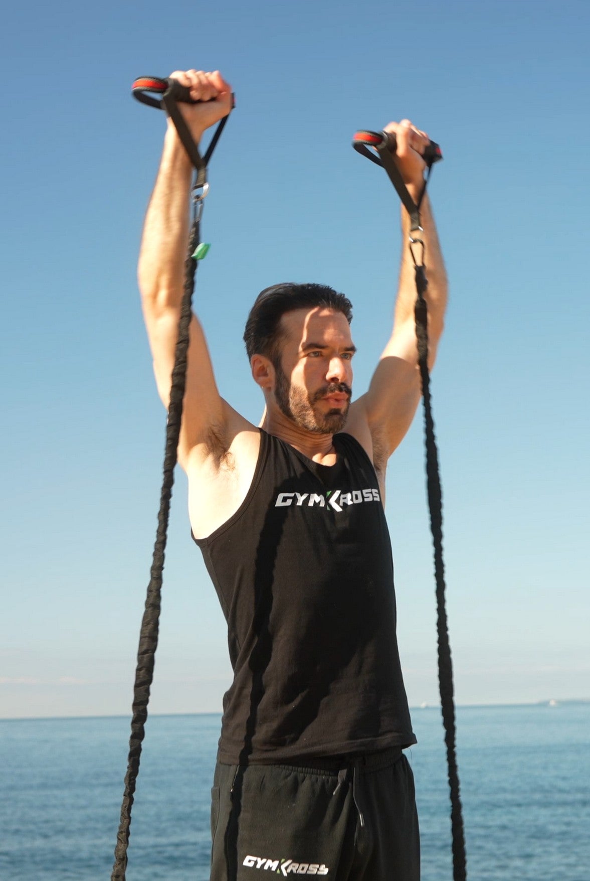 Un homme portant un débardeur Gymkross et un pantalon noir s&#39;entraîne avec des bandes de résistance en plein air près de la mer sous un ciel bleu clair.