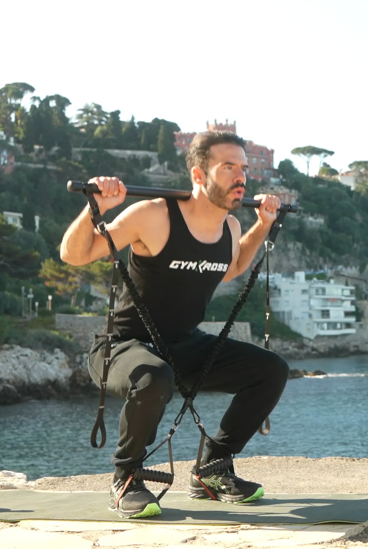 Homme réalisant des squats avec une barre et des sangles de résistance GymKross, vêtu d&#39;un débardeur noir et de baskets, en plein air sur un pont en pierre, concentré sur sa routine de fitness pour renforcer les jambes.